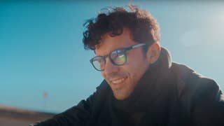 A man wears smart glasses against a blue sky and smiles to himself.